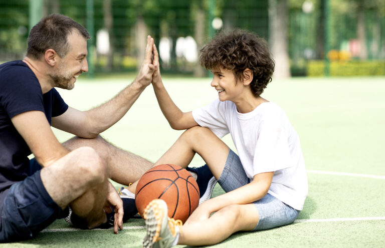 Portrait of dad and son giving high five to each other, sitting at the playground with a basketball